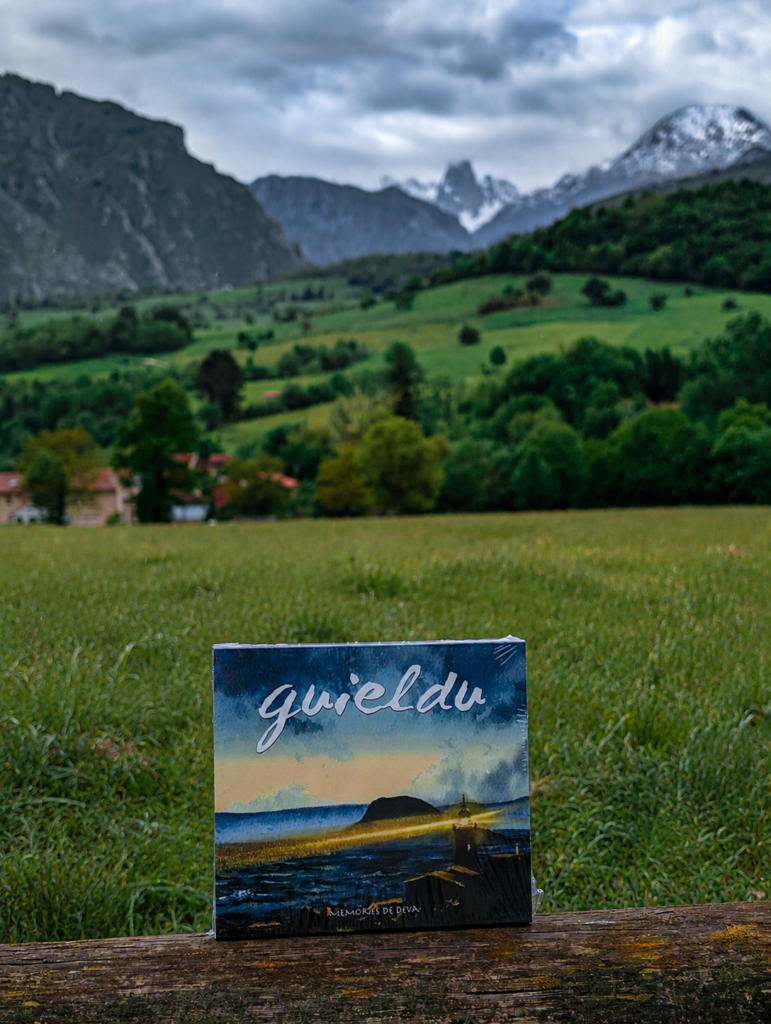 El CD del álbum «Memories de Deva» de Guieldu apoyado sobre una barandilla de madera, con los prados de Cabrales y el macizo de los Urrieles (Pico Urriellu) al fondo.
